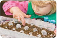 blonde girl wearing pink shirt green apron planting seeds in egg carton