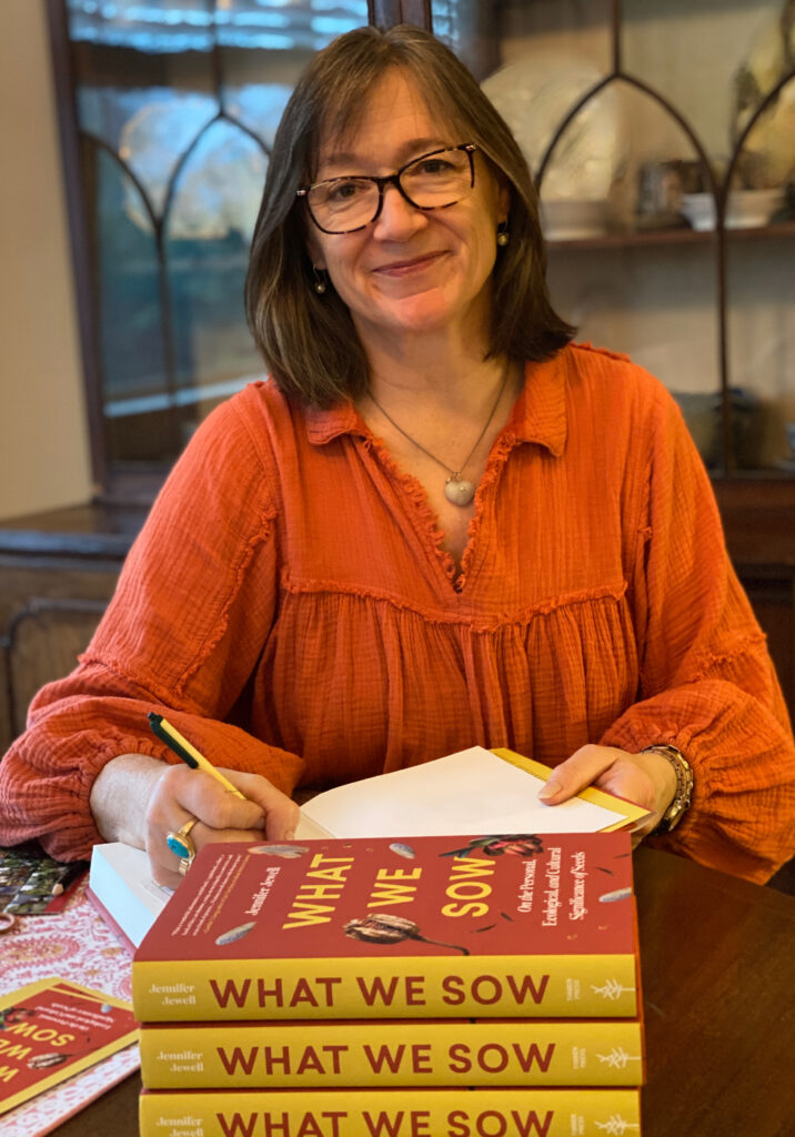 Photo of Jennifer Jewell - she is a white woman who wears black rimmed glasses, has shoulder length brown hair, is wearing an orange/rust color shirt and is sitting in front of three copies of her book "what we sow"