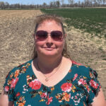 white woman with brown hair pulled back, wearing sunglasses and a blue blouse with red and orange flowers. woman is standing in a field - left side is tilled; right side is not and is green