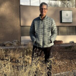 white woman with short salt and pepper hair wearing black jeans and silver jacket standing outside behind cattails and in front of a brown building with the El Paso County log on on the wall.