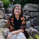 white woman with shoulder length dark brown hair wearing glasses, khaki pants and a brown shirt with pink and orange flowers. she is sitting down in a rock garden