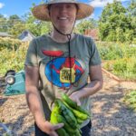 white woman wearing a straw hat, short brown haird, green t-shirt with "spicy chef" graphic that is a cartoon red "devil" in a yellow apron holding a red pepper on a pitchfork. The woman is holding about seven green peppers and she is standing in a small community garden.